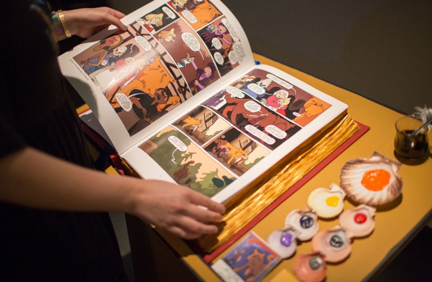 Photo by Bridget Badore, a display of shells with different colored paints in each one, next to light skinned hands holding an open comic book depicting characters with a large orange cat.