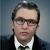 A white figure with glasses and a suit looks at the camera.