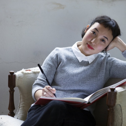 A portrait depicts an Asian woman with medium light skin, black hair, red lipstick, and a gray sweater sitting in an ivory chair against a gray wall. She looks at the viewer while setting a pen to the open book in her lap.