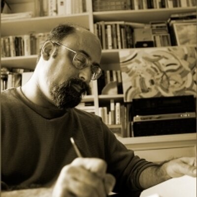 A sepia-toned photograph shows a bearded figure with glasses writing on paper. Behind the figure is a bookcase stacked with books and art.