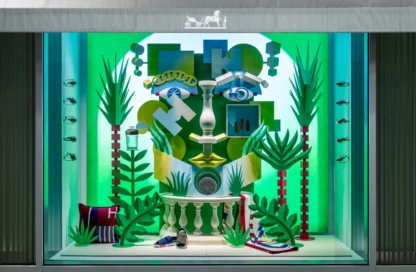 An Hermes window display. Inside, a brightly colored green display with fake trees and objects made up to resemble a face.