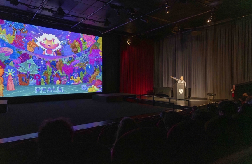 Photograph of an auditorium. A student stands in front of a podium on the right. A large screen with a psychedelic purple, blue, and pink illustration appears on a screen in the center of the stage.