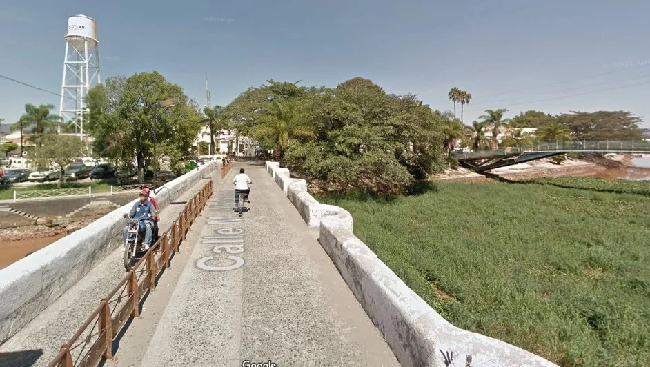 Google Maps screenshot of a town, from the viewpoint of a bridge. To the left, a water tower stands in the distance. To the right, a polluted river is overgrown with vegetation.