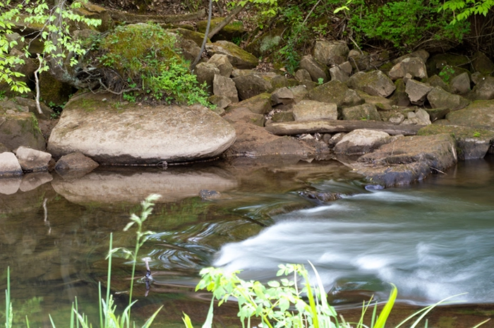 A photograph of a creek in the woods. Rocks are piled across the creek, creating a small waterfall. On the banks of the creek are large boulders that create a hill that leads up into the woods.