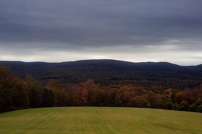 A photograph of rolling mountains in Pennsylvania. The clouds are dark, casting a blue shadow over the mountains. The trees in front of the mountains are bright red and orange.