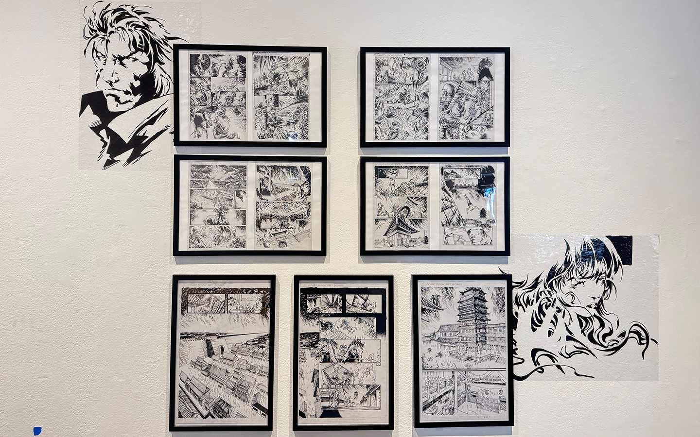 It's a photo of a wall, exhibiting seven black frames with two B&W pin-up vinyls on upper left and bottom right corners.