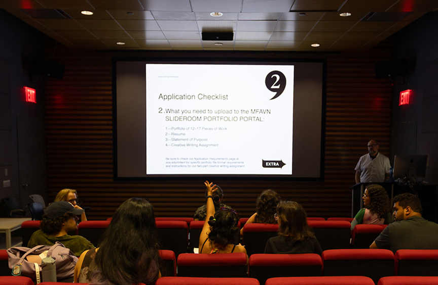 Wide shot of an auditorium with students sitting facing a large projector screen that reads