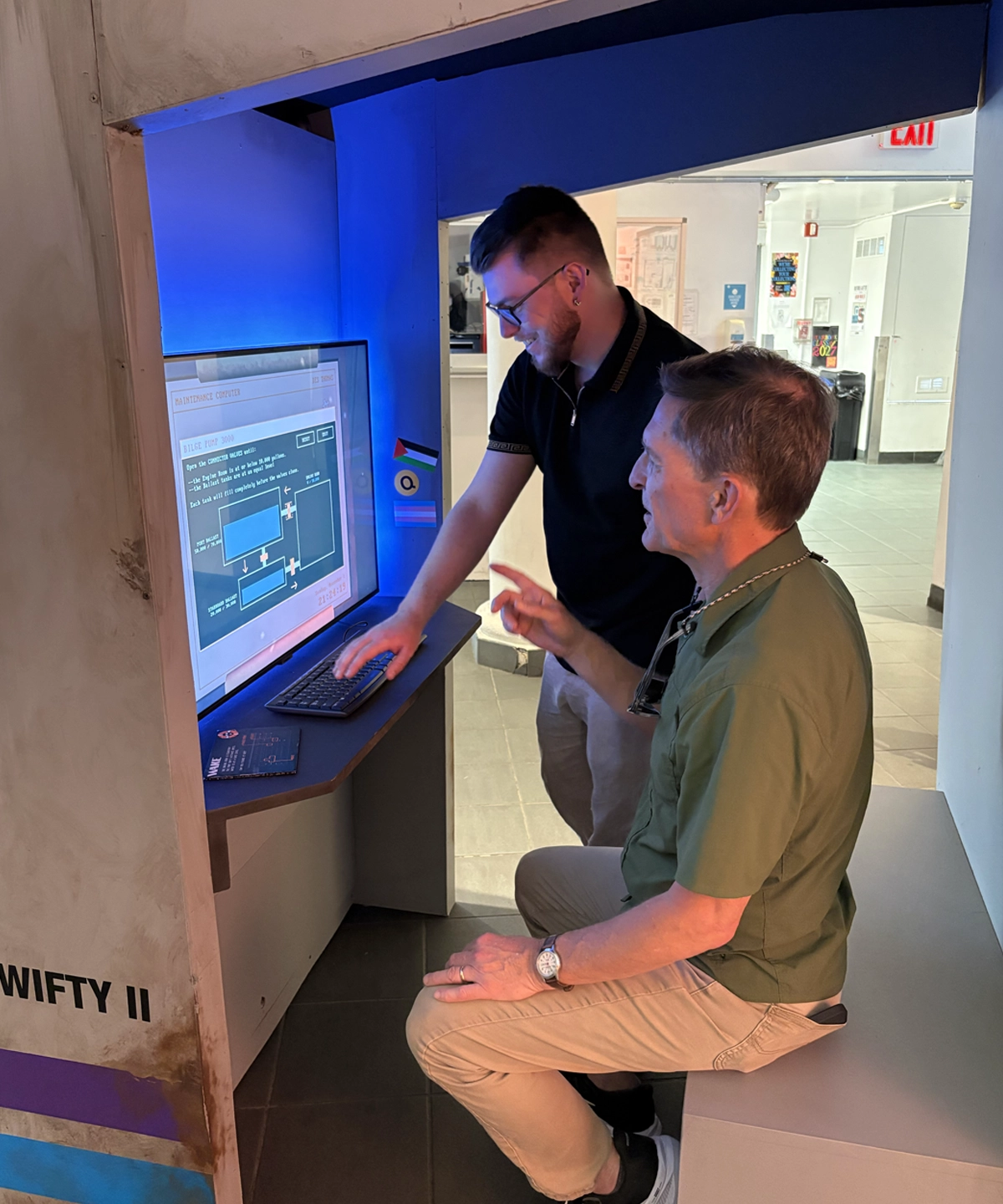 Two people sit in the booth playing the demo game on the screen inside.
