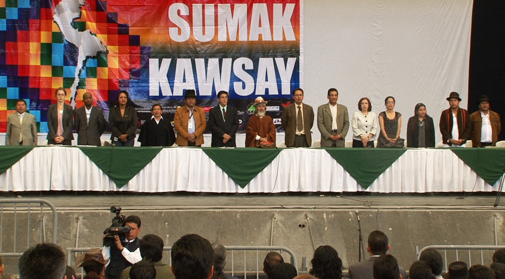 A row of officials and community representatives stands behind a long draped table on stage, beneath a large colorful banner reading 