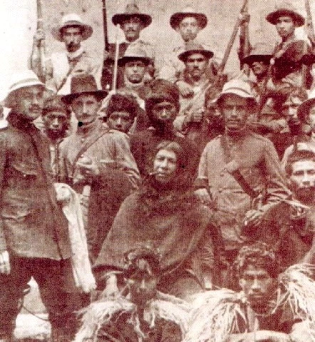 Indigenous Colombian leader Quintín Lame stands at the center of the frame, wrapped in a poncho and smoking, surrounded by armed men in military-style dress and hats. The sepia tone and tense grouping capture the moment of his arrest, a quiet but charged image of resistance confronted by state force.