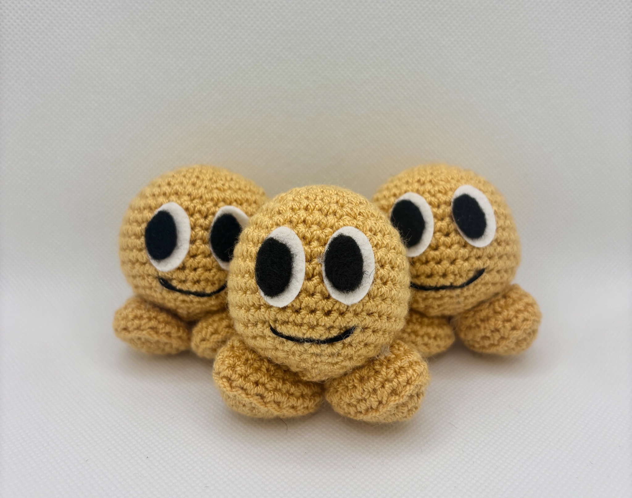 Three matching crocheted buñuelo dolls sit side by side against a plain white background. Each has a round golden-yellow head, big black-and-white felt eyes, a stitched smiling mouth, and short, rounded legs peeking out beneath.