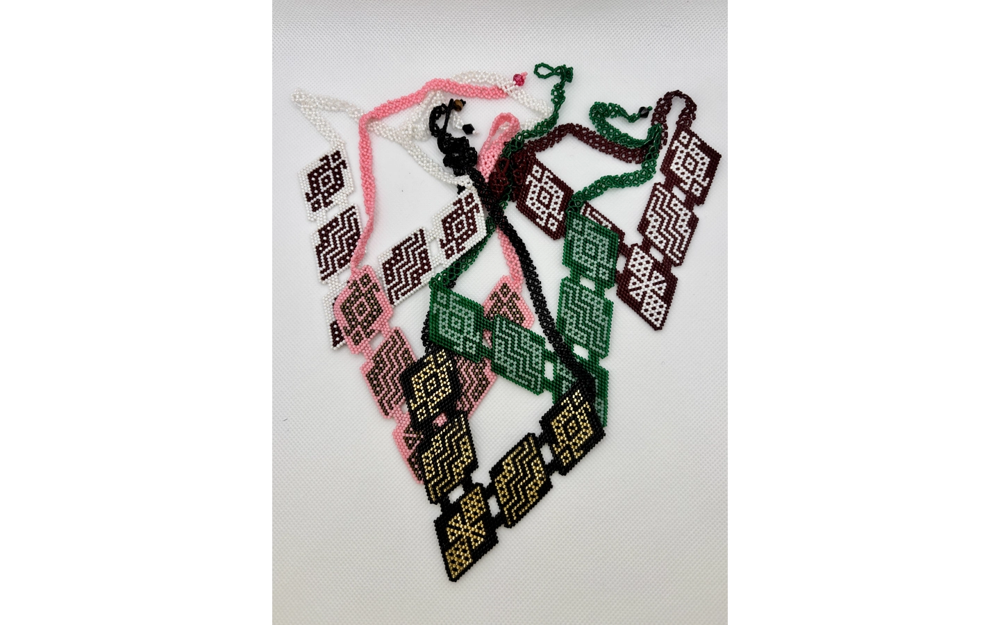 Several beaded necklaces, made in Inga and Kamëntsá beadwork traditions, overlap in a tangled pile against a white background. Each strand features diamond-shaped panels in a different color palette — maroon and white, pink and brown, green and white, black and gold — with geometric patterns that repeat across each piece with slight variations.