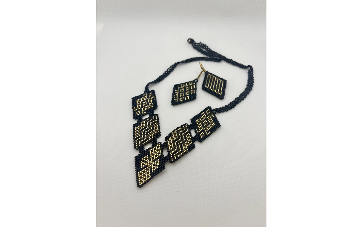 A beaded necklace and matching earrings, made using Inga and Kamëntsá beadwork traditions, lie against a plain white background. Woven in black beads with intricate geometric patterns picked out in gold, diamond-shaped panels graduate in size down the necklace's center, echoing the smaller diamond earrings beside them.