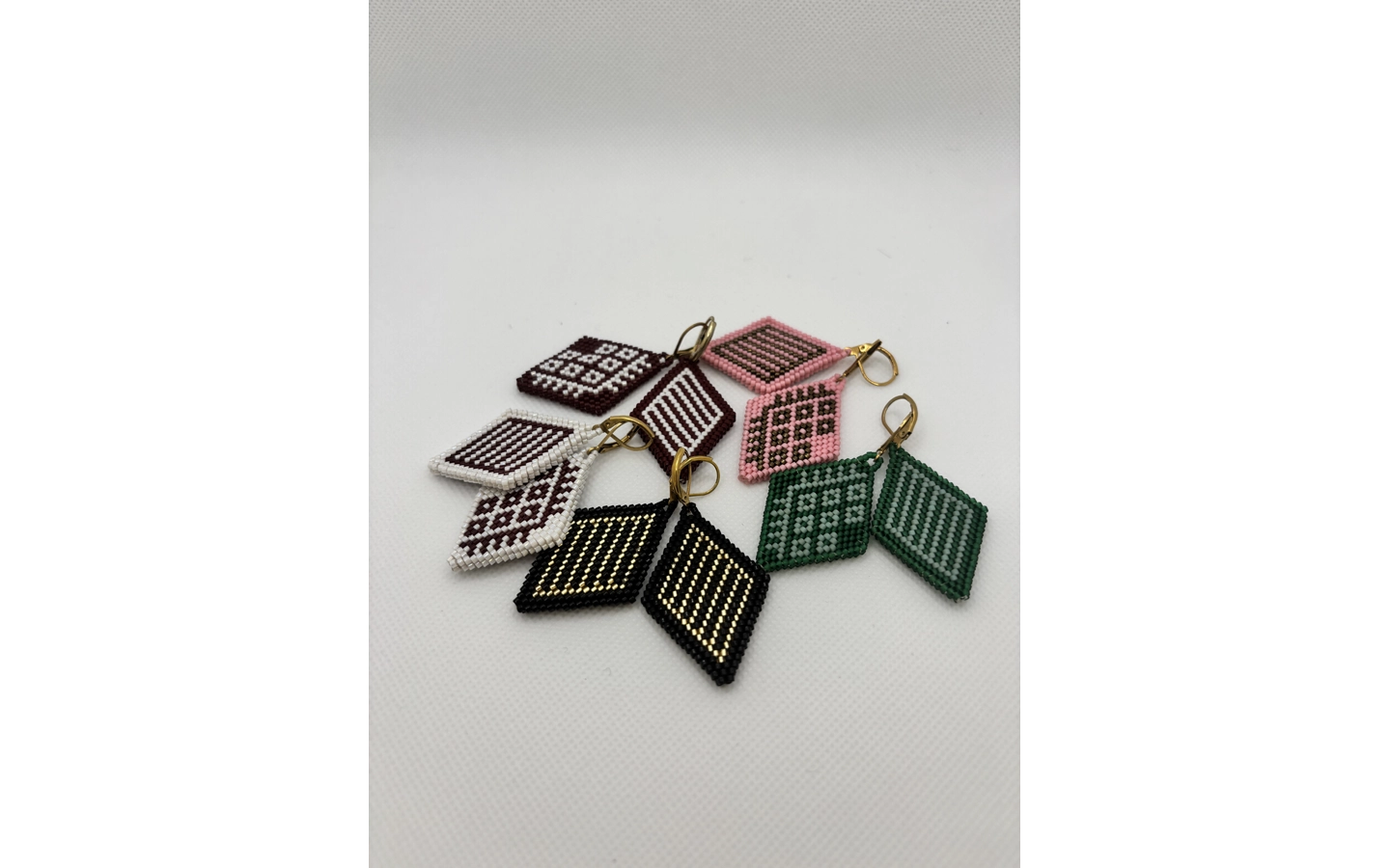 Five pairs of diamond-shaped beaded earrings, crafted in Inga and Kamëntsá beadwork style, are arranged loosely on a white surface. Each pair appears in a different two-tone combination, including maroon and white, pink and brown, black and gold, and green and white, with small gold hooks and consistent geometric motifs unifying the set.