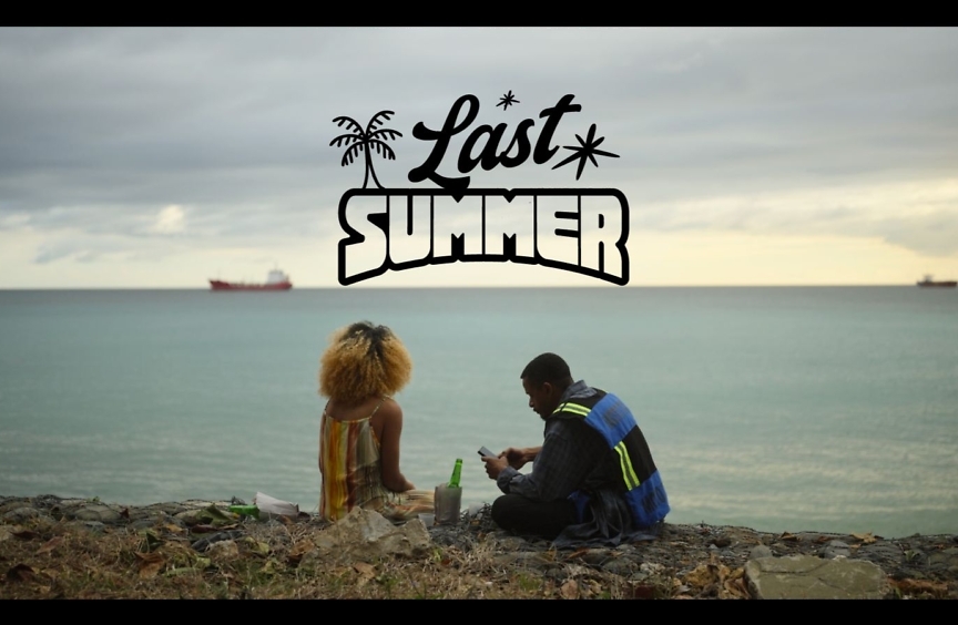 Last Summer title card: a woman and a man sit together on a rocky shoreline looking out over the sea, with cargo ships on the horizon.